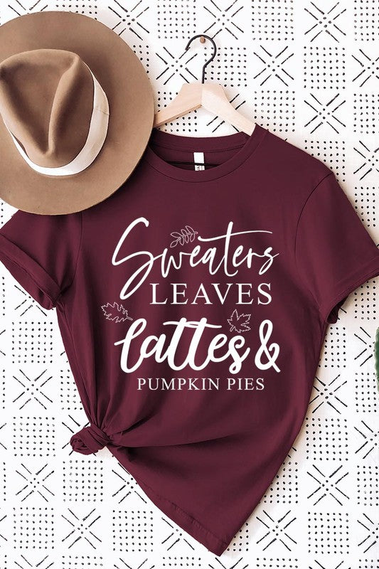 A white "Sweaters,Leaves Graphic Tee" hangs on a wooden hanger, featuring the text "Sweaters, Leaves, Lattes & Pumpkin Pies." In the background, a cozy knitted sweater adds to the inviting scene. This unisex T-shirt blends seasonal charm with everyday comfort.