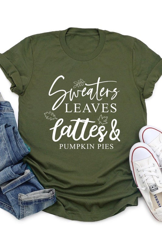 A white "Sweaters,Leaves Graphic Tee" hangs on a wooden hanger, featuring the text "Sweaters, Leaves, Lattes & Pumpkin Pies." In the background, a cozy knitted sweater adds to the inviting scene. This unisex T-shirt blends seasonal charm with everyday comfort.