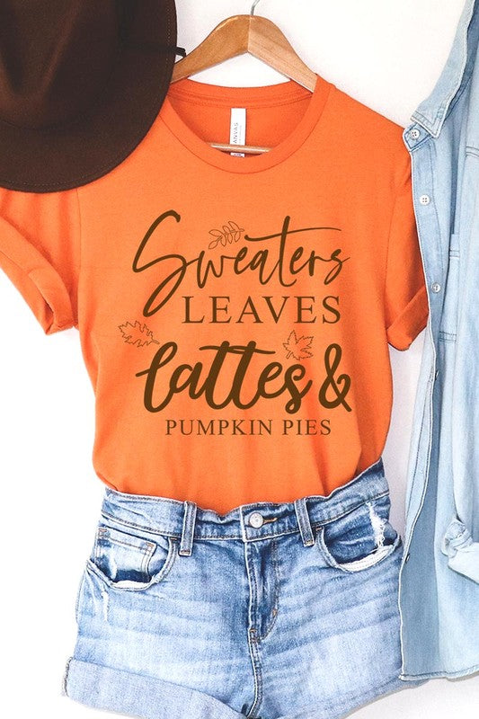 A white "Sweaters,Leaves Graphic Tee" hangs on a wooden hanger, featuring the text "Sweaters, Leaves, Lattes & Pumpkin Pies." In the background, a cozy knitted sweater adds to the inviting scene. This unisex T-shirt blends seasonal charm with everyday comfort.