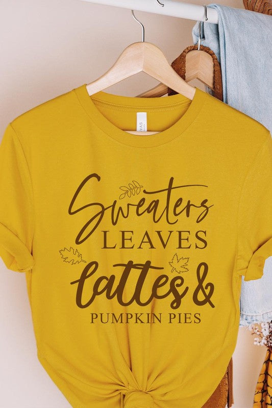 A white "Sweaters,Leaves Graphic Tee" hangs on a wooden hanger, featuring the text "Sweaters, Leaves, Lattes & Pumpkin Pies." In the background, a cozy knitted sweater adds to the inviting scene. This unisex T-shirt blends seasonal charm with everyday comfort.