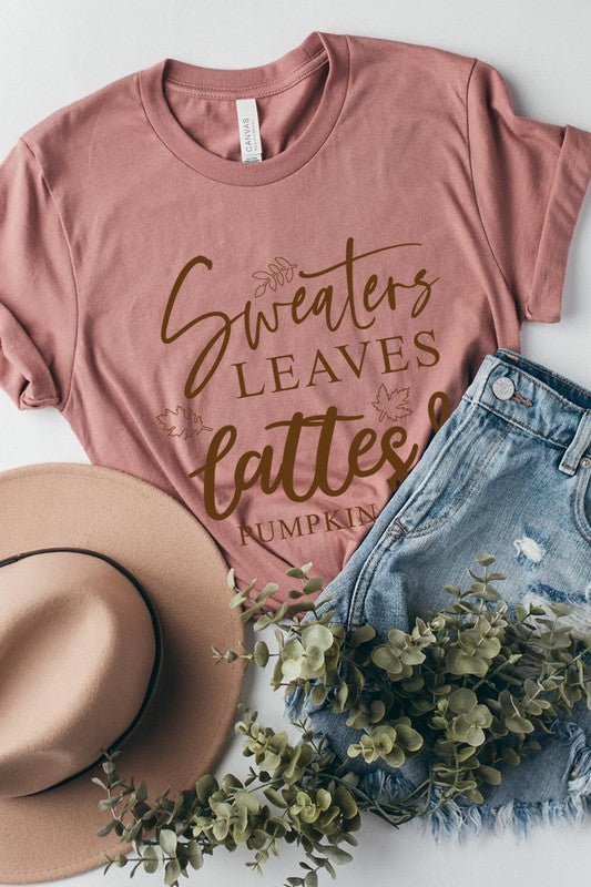 A white "Sweaters,Leaves Graphic Tee" hangs on a wooden hanger, featuring the text "Sweaters, Leaves, Lattes & Pumpkin Pies." In the background, a cozy knitted sweater adds to the inviting scene. This unisex T-shirt blends seasonal charm with everyday comfort.