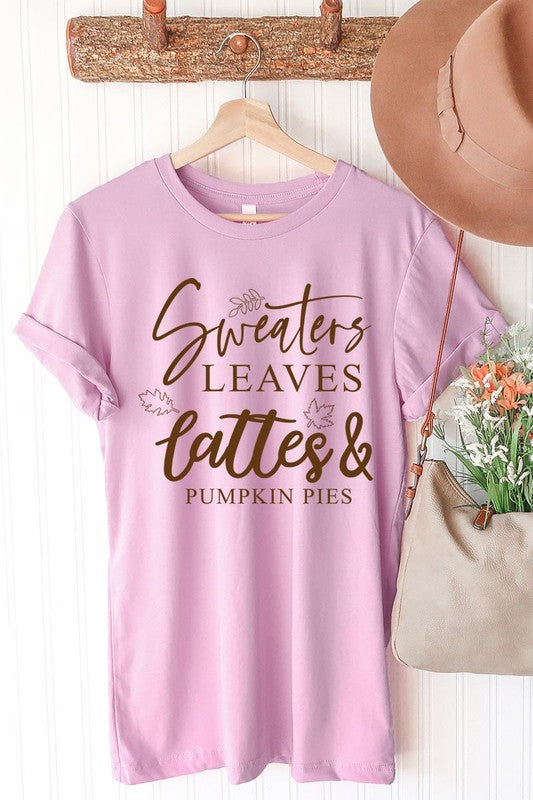 A white "Sweaters,Leaves Graphic Tee" hangs on a wooden hanger, featuring the text "Sweaters, Leaves, Lattes & Pumpkin Pies." In the background, a cozy knitted sweater adds to the inviting scene. This unisex T-shirt blends seasonal charm with everyday comfort.