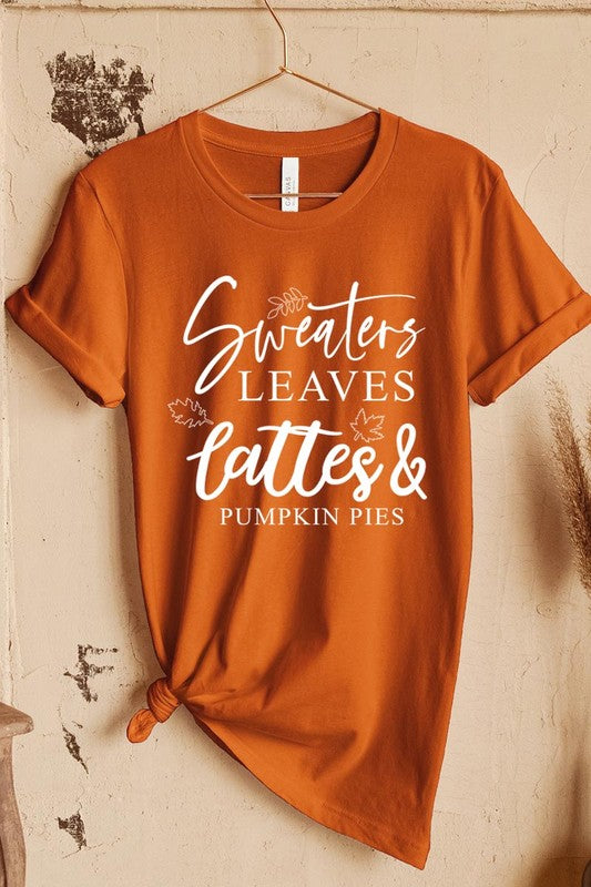 A white "Sweaters,Leaves Graphic Tee" hangs on a wooden hanger, featuring the text "Sweaters, Leaves, Lattes & Pumpkin Pies." In the background, a cozy knitted sweater adds to the inviting scene. This unisex T-shirt blends seasonal charm with everyday comfort.