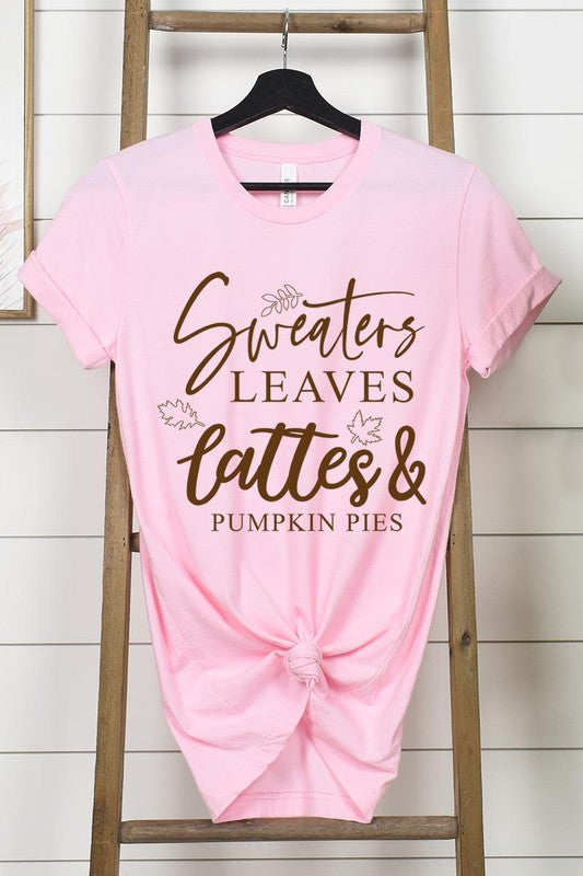 A white "Sweaters,Leaves Graphic Tee" hangs on a wooden hanger, featuring the text "Sweaters, Leaves, Lattes & Pumpkin Pies." In the background, a cozy knitted sweater adds to the inviting scene. This unisex T-shirt blends seasonal charm with everyday comfort.