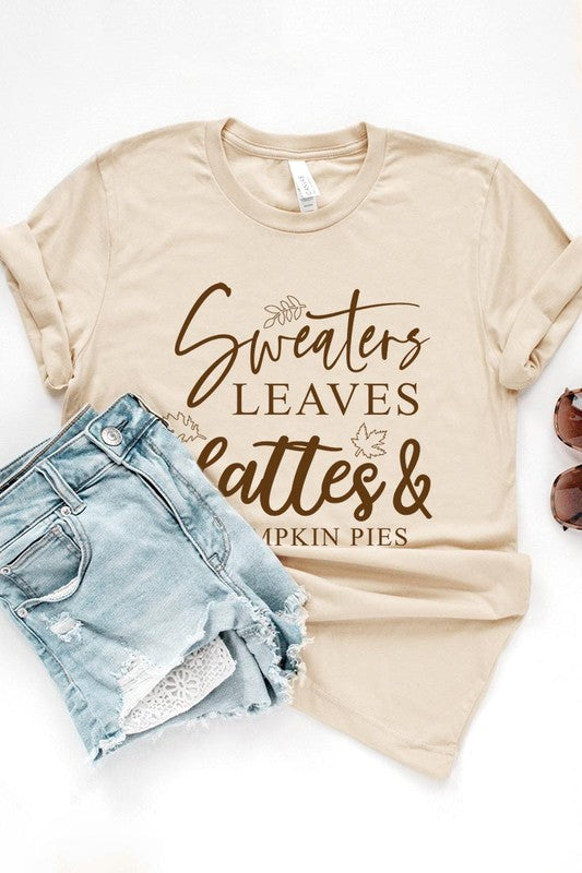 A white "Sweaters,Leaves Graphic Tee" hangs on a wooden hanger, featuring the text "Sweaters, Leaves, Lattes & Pumpkin Pies." In the background, a cozy knitted sweater adds to the inviting scene. This unisex T-shirt blends seasonal charm with everyday comfort.