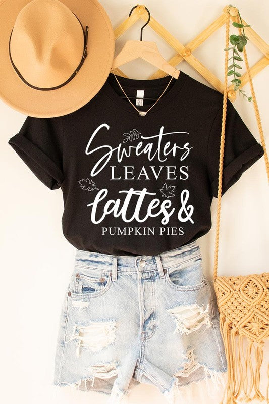 A white "Sweaters,Leaves Graphic Tee" hangs on a wooden hanger, featuring the text "Sweaters, Leaves, Lattes & Pumpkin Pies." In the background, a cozy knitted sweater adds to the inviting scene. This unisex T-shirt blends seasonal charm with everyday comfort.