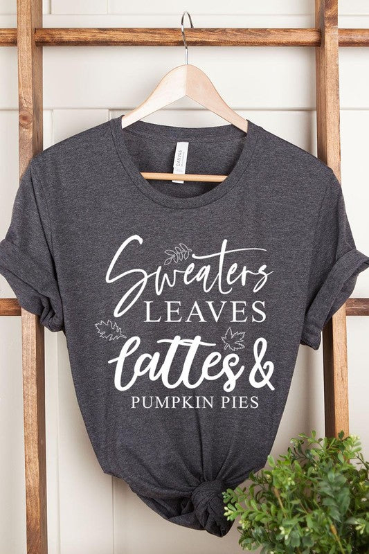 A white "Sweaters,Leaves Graphic Tee" hangs on a wooden hanger, featuring the text "Sweaters, Leaves, Lattes & Pumpkin Pies." In the background, a cozy knitted sweater adds to the inviting scene. This unisex T-shirt blends seasonal charm with everyday comfort.