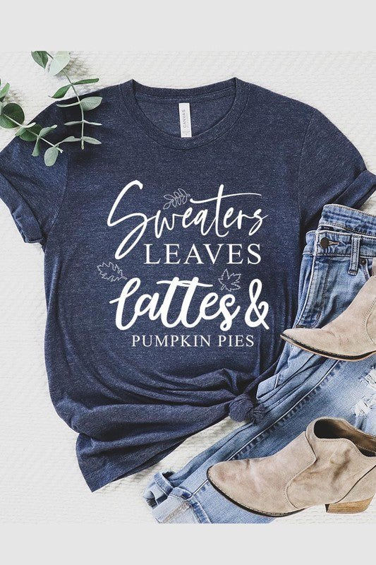 A white "Sweaters,Leaves Graphic Tee" hangs on a wooden hanger, featuring the text "Sweaters, Leaves, Lattes & Pumpkin Pies." In the background, a cozy knitted sweater adds to the inviting scene. This unisex T-shirt blends seasonal charm with everyday comfort.