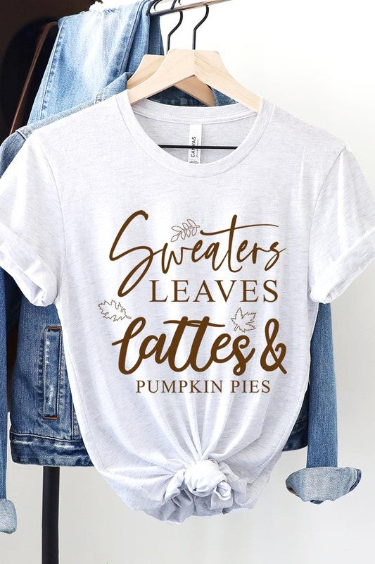 A white "Sweaters,Leaves Graphic Tee" hangs on a wooden hanger, featuring the text "Sweaters, Leaves, Lattes & Pumpkin Pies." In the background, a cozy knitted sweater adds to the inviting scene. This unisex T-shirt blends seasonal charm with everyday comfort.