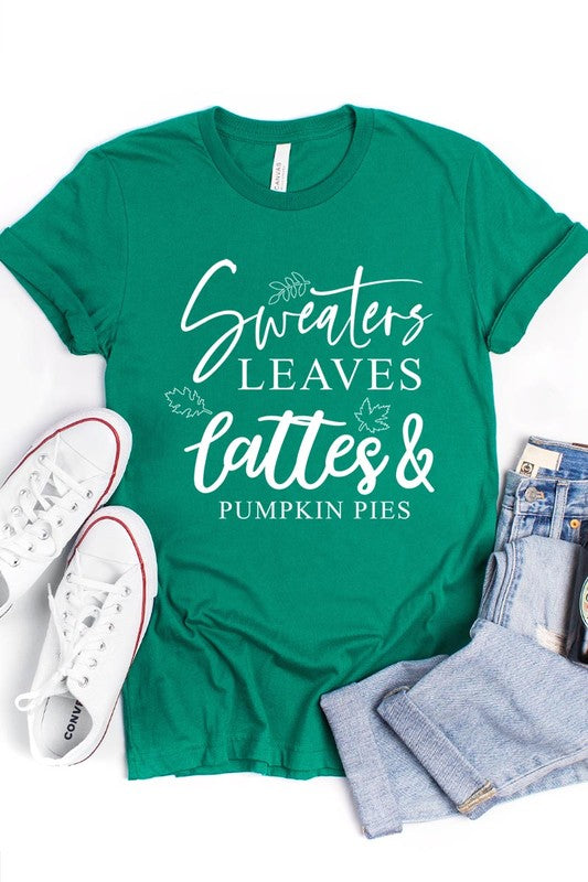 A white "Sweaters,Leaves Graphic Tee" hangs on a wooden hanger, featuring the text "Sweaters, Leaves, Lattes & Pumpkin Pies." In the background, a cozy knitted sweater adds to the inviting scene. This unisex T-shirt blends seasonal charm with everyday comfort.