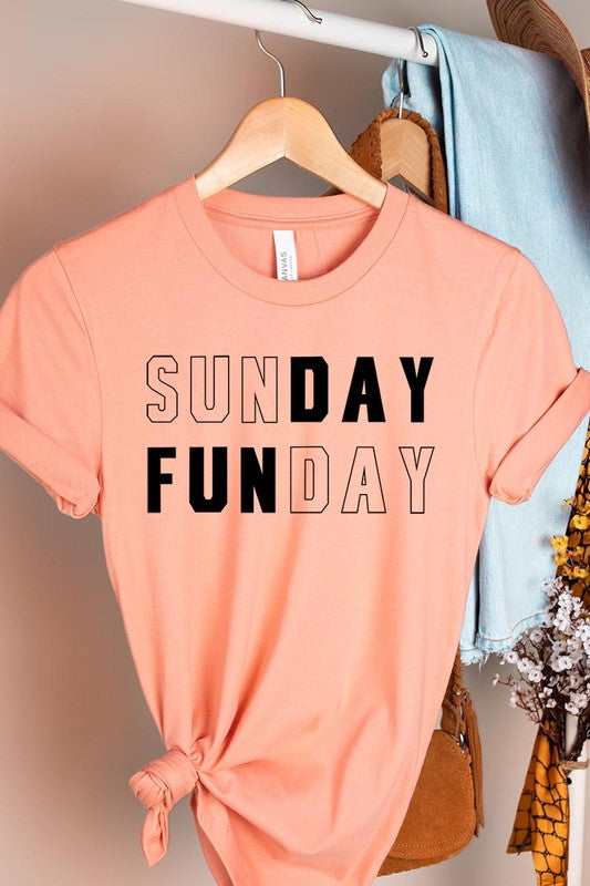 A light pink Sunday Funday Graphic Tee featuring the "SUNDAY FUNDAY" print is hanging on a wooden hanger next to a tan hat and a beige bag filled with flowers. This unisex crew neck short sleeve tee is ideal for family group uniforms.