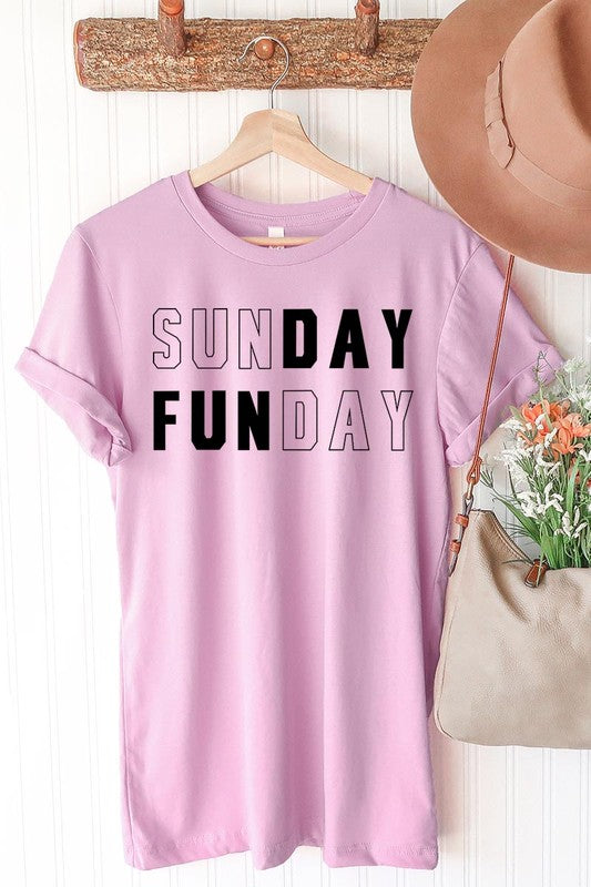 A light pink Sunday Funday Graphic Tee featuring the "SUNDAY FUNDAY" print is hanging on a wooden hanger next to a tan hat and a beige bag filled with flowers. This unisex crew neck short sleeve tee is ideal for family group uniforms.