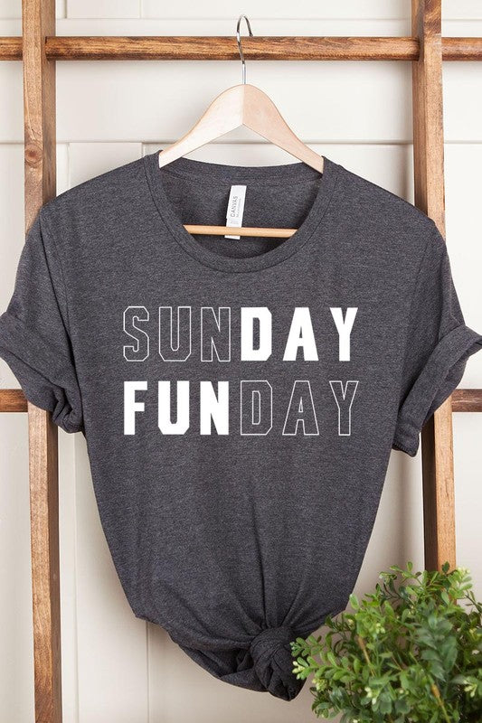 A light pink Sunday Funday Graphic Tee featuring the "SUNDAY FUNDAY" print is hanging on a wooden hanger next to a tan hat and a beige bag filled with flowers. This unisex crew neck short sleeve tee is ideal for family group uniforms.