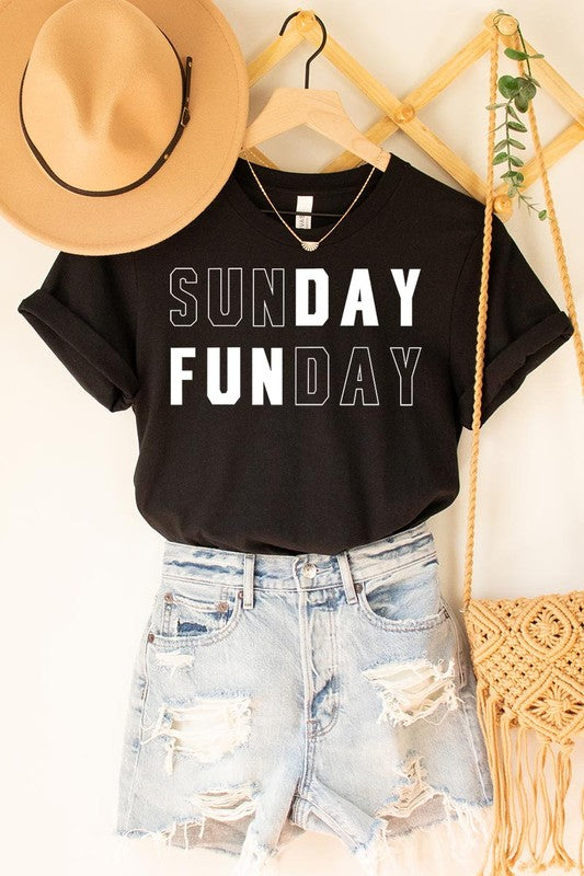 A light pink Sunday Funday Graphic Tee featuring the "SUNDAY FUNDAY" print is hanging on a wooden hanger next to a tan hat and a beige bag filled with flowers. This unisex crew neck short sleeve tee is ideal for family group uniforms.