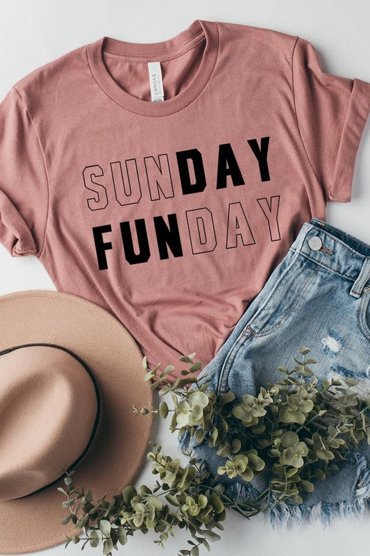 A light pink Sunday Funday Graphic Tee featuring the "SUNDAY FUNDAY" print is hanging on a wooden hanger next to a tan hat and a beige bag filled with flowers. This unisex crew neck short sleeve tee is ideal for family group uniforms.