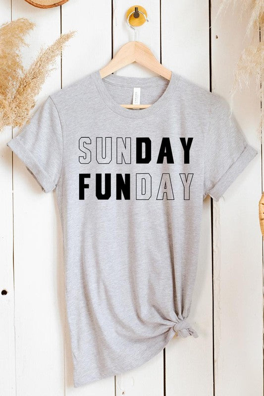 A light pink Sunday Funday Graphic Tee featuring the "SUNDAY FUNDAY" print is hanging on a wooden hanger next to a tan hat and a beige bag filled with flowers. This unisex crew neck short sleeve tee is ideal for family group uniforms.