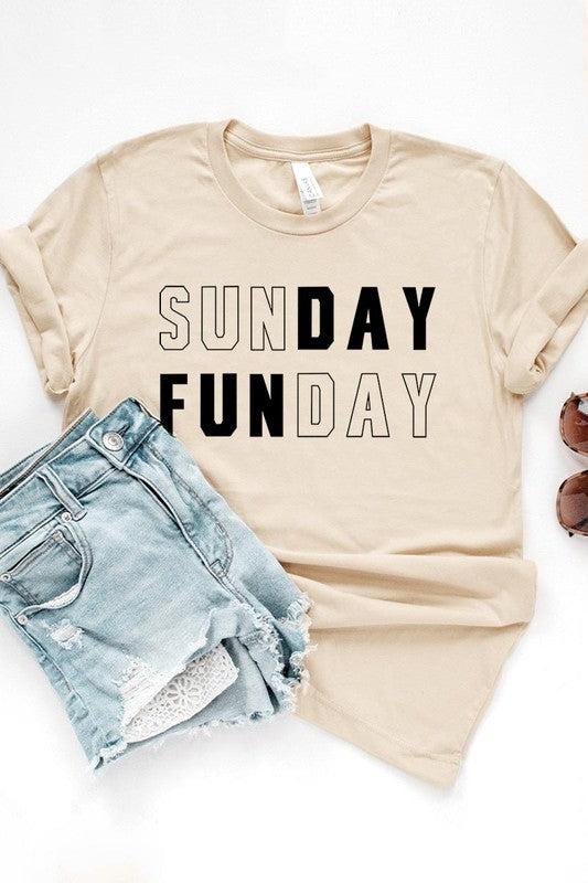 A light pink Sunday Funday Graphic Tee featuring the "SUNDAY FUNDAY" print is hanging on a wooden hanger next to a tan hat and a beige bag filled with flowers. This unisex crew neck short sleeve tee is ideal for family group uniforms.