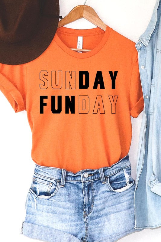 A light pink Sunday Funday Graphic Tee featuring the "SUNDAY FUNDAY" print is hanging on a wooden hanger next to a tan hat and a beige bag filled with flowers. This unisex crew neck short sleeve tee is ideal for family group uniforms.