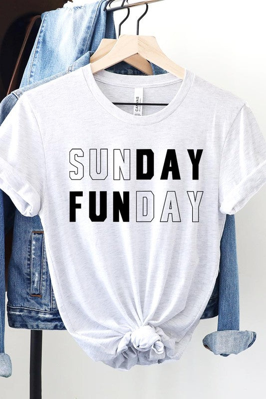 A light pink Sunday Funday Graphic Tee featuring the "SUNDAY FUNDAY" print is hanging on a wooden hanger next to a tan hat and a beige bag filled with flowers. This unisex crew neck short sleeve tee is ideal for family group uniforms.