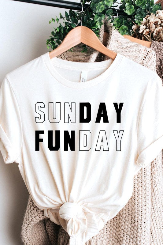 A light pink Sunday Funday Graphic Tee featuring the "SUNDAY FUNDAY" print is hanging on a wooden hanger next to a tan hat and a beige bag filled with flowers. This unisex crew neck short sleeve tee is ideal for family group uniforms.