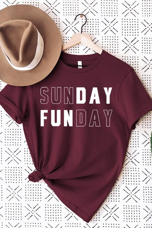 A light pink Sunday Funday Graphic Tee featuring the "SUNDAY FUNDAY" print is hanging on a wooden hanger next to a tan hat and a beige bag filled with flowers. This unisex crew neck short sleeve tee is ideal for family group uniforms.