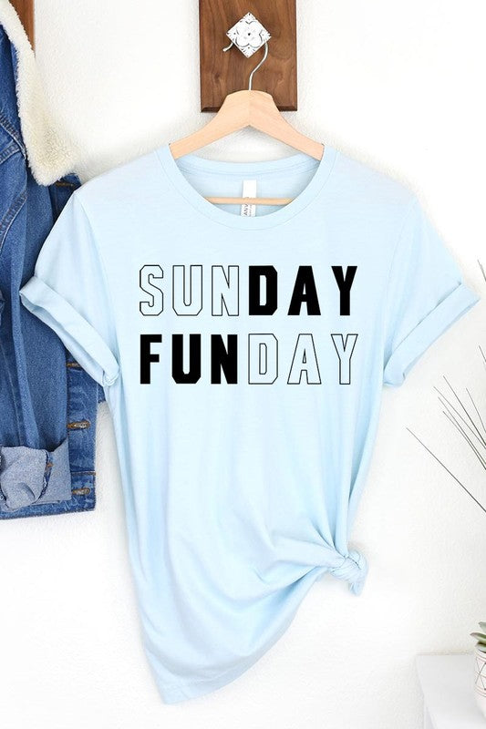 A light pink Sunday Funday Graphic Tee featuring the "SUNDAY FUNDAY" print is hanging on a wooden hanger next to a tan hat and a beige bag filled with flowers. This unisex crew neck short sleeve tee is ideal for family group uniforms.