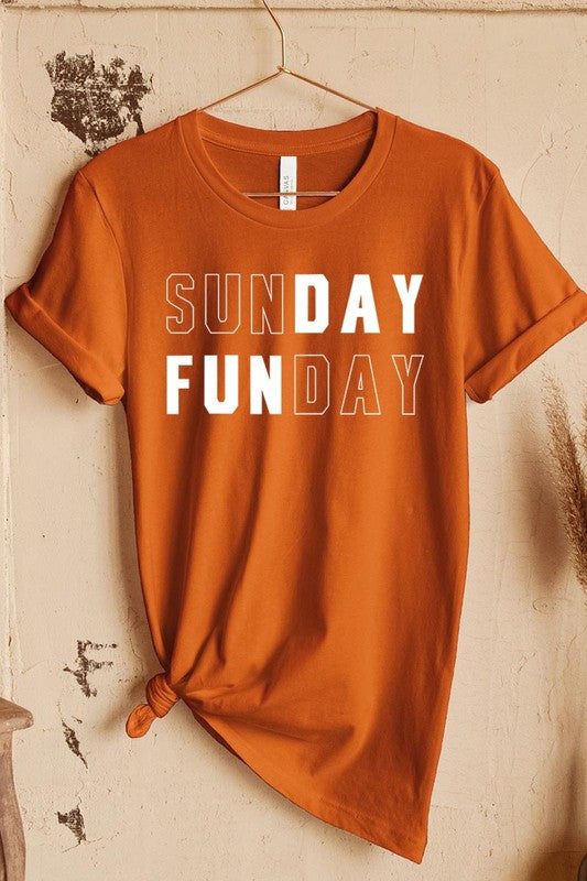 A light pink Sunday Funday Graphic Tee featuring the "SUNDAY FUNDAY" print is hanging on a wooden hanger next to a tan hat and a beige bag filled with flowers. This unisex crew neck short sleeve tee is ideal for family group uniforms.