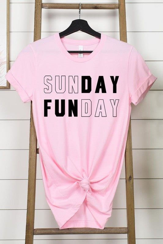 A light pink Sunday Funday Graphic Tee featuring the "SUNDAY FUNDAY" print is hanging on a wooden hanger next to a tan hat and a beige bag filled with flowers. This unisex crew neck short sleeve tee is ideal for family group uniforms.