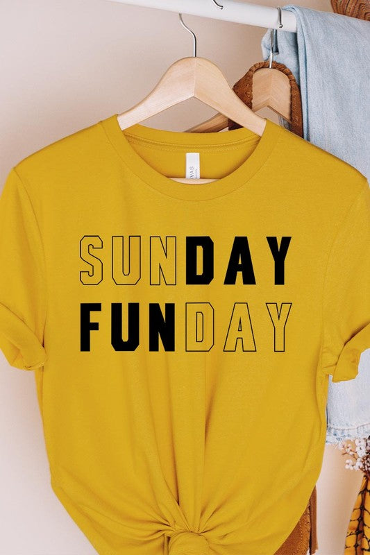 A light pink Sunday Funday Graphic Tee featuring the "SUNDAY FUNDAY" print is hanging on a wooden hanger next to a tan hat and a beige bag filled with flowers. This unisex crew neck short sleeve tee is ideal for family group uniforms.