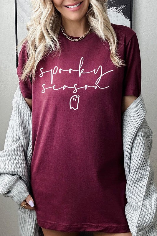 Person sitting on a stool, wearing the "Spooky Season Graphic Tee" that features the text "Spooky Season" and a ghost graphic, holding a book titled "Spells & Potions" in front of their face. Black bat decorations adorn the wall in the background.