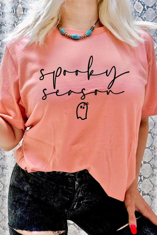 Person sitting on a stool, wearing the "Spooky Season Graphic Tee" that features the text "Spooky Season" and a ghost graphic, holding a book titled "Spells & Potions" in front of their face. Black bat decorations adorn the wall in the background.