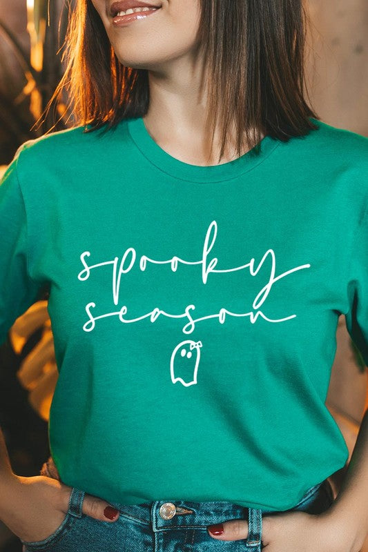 Person sitting on a stool, wearing the "Spooky Season Graphic Tee" that features the text "Spooky Season" and a ghost graphic, holding a book titled "Spells & Potions" in front of their face. Black bat decorations adorn the wall in the background.