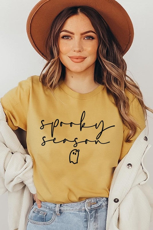 Person sitting on a stool, wearing the "Spooky Season Graphic Tee" that features the text "Spooky Season" and a ghost graphic, holding a book titled "Spells & Potions" in front of their face. Black bat decorations adorn the wall in the background.