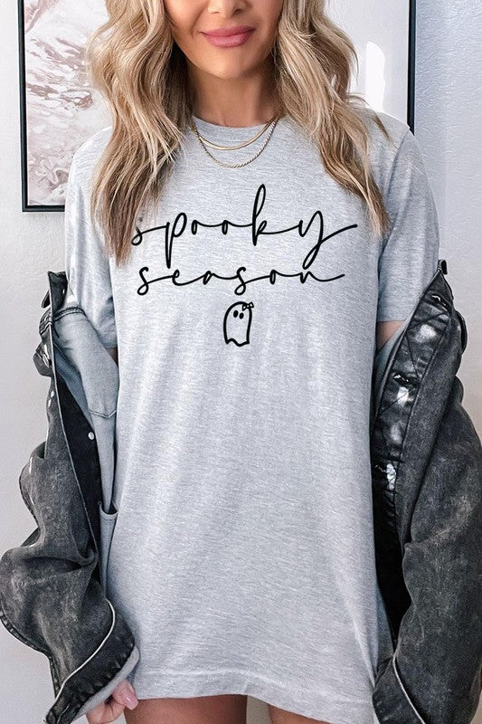 Person sitting on a stool, wearing the "Spooky Season Graphic Tee" that features the text "Spooky Season" and a ghost graphic, holding a book titled "Spells & Potions" in front of their face. Black bat decorations adorn the wall in the background.