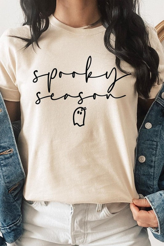 Person sitting on a stool, wearing the "Spooky Season Graphic Tee" that features the text "Spooky Season" and a ghost graphic, holding a book titled "Spells & Potions" in front of their face. Black bat decorations adorn the wall in the background.