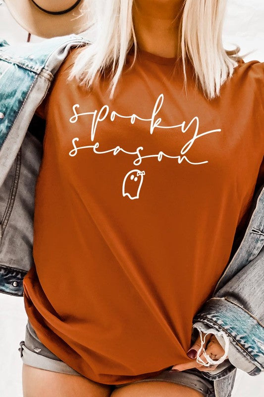 Person sitting on a stool, wearing the "Spooky Season Graphic Tee" that features the text "Spooky Season" and a ghost graphic, holding a book titled "Spells & Potions" in front of their face. Black bat decorations adorn the wall in the background.