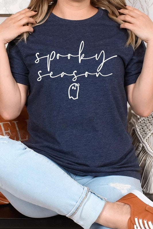 Person sitting on a stool, wearing the "Spooky Season Graphic Tee" that features the text "Spooky Season" and a ghost graphic, holding a book titled "Spells & Potions" in front of their face. Black bat decorations adorn the wall in the background.