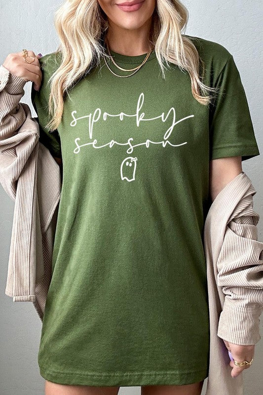 Person sitting on a stool, wearing the "Spooky Season Graphic Tee" that features the text "Spooky Season" and a ghost graphic, holding a book titled "Spells & Potions" in front of their face. Black bat decorations adorn the wall in the background.