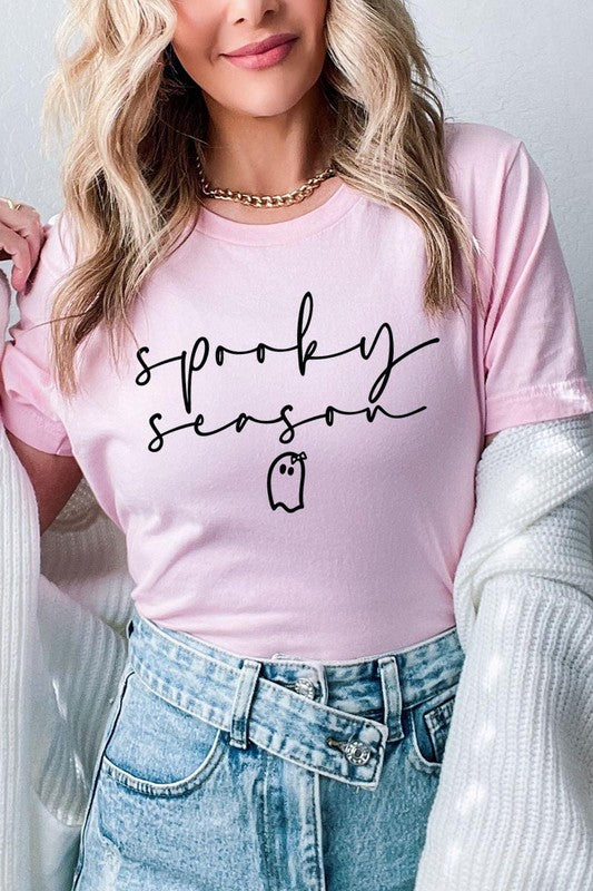 Person sitting on a stool, wearing the "Spooky Season Graphic Tee" that features the text "Spooky Season" and a ghost graphic, holding a book titled "Spells & Potions" in front of their face. Black bat decorations adorn the wall in the background.