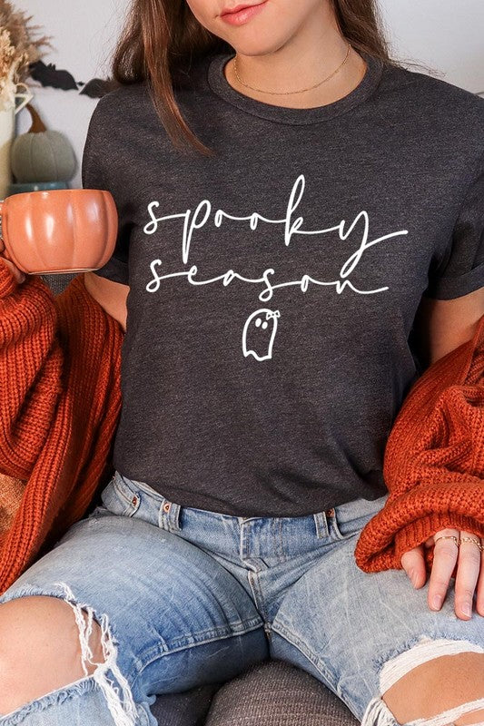 Person sitting on a stool, wearing the "Spooky Season Graphic Tee" that features the text "Spooky Season" and a ghost graphic, holding a book titled "Spells & Potions" in front of their face. Black bat decorations adorn the wall in the background.