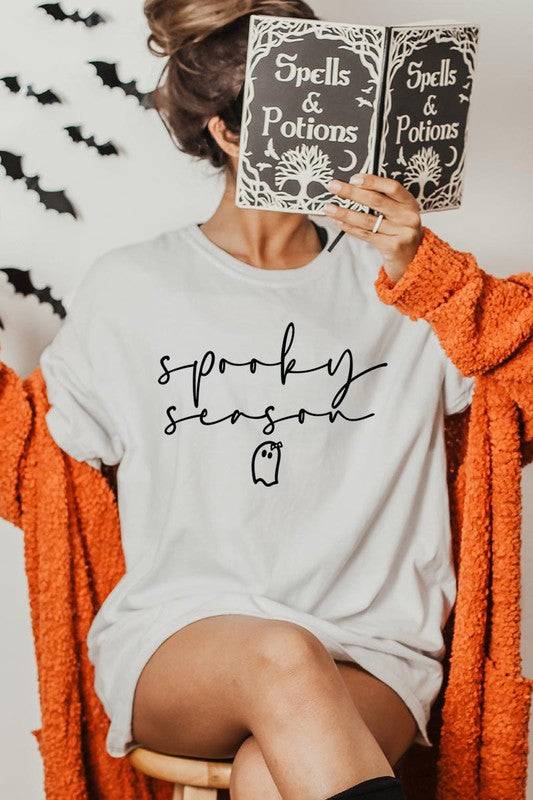 Person sitting on a stool, wearing the "Spooky Season Graphic Tee" that features the text "Spooky Season" and a ghost graphic, holding a book titled "Spells & Potions" in front of their face. Black bat decorations adorn the wall in the background.