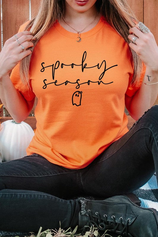 Person sitting on a stool, wearing the "Spooky Season Graphic Tee" that features the text "Spooky Season" and a ghost graphic, holding a book titled "Spells & Potions" in front of their face. Black bat decorations adorn the wall in the background.