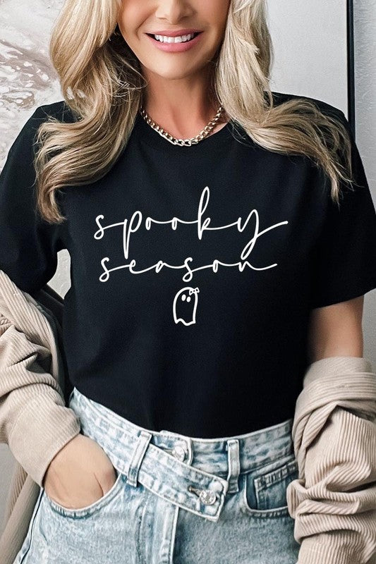 Person sitting on a stool, wearing the "Spooky Season Graphic Tee" that features the text "Spooky Season" and a ghost graphic, holding a book titled "Spells & Potions" in front of their face. Black bat decorations adorn the wall in the background.