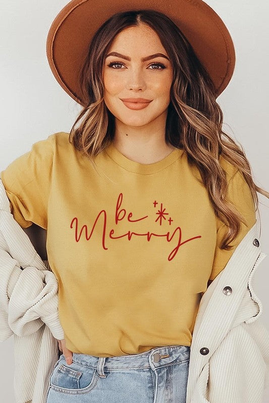 A person wearing the stylish and high-quality Be Merry Graphic Tee, featuring a decorative design with light-colored fabric and bold red text that reads "Be Merry." The unisex crew neck shirt is perfectly paired with them holding a red and black plaid garment.