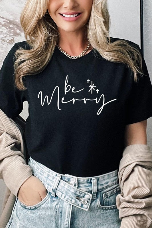 A person wearing the stylish and high-quality Be Merry Graphic Tee, featuring a decorative design with light-colored fabric and bold red text that reads "Be Merry." The unisex crew neck shirt is perfectly paired with them holding a red and black plaid garment.