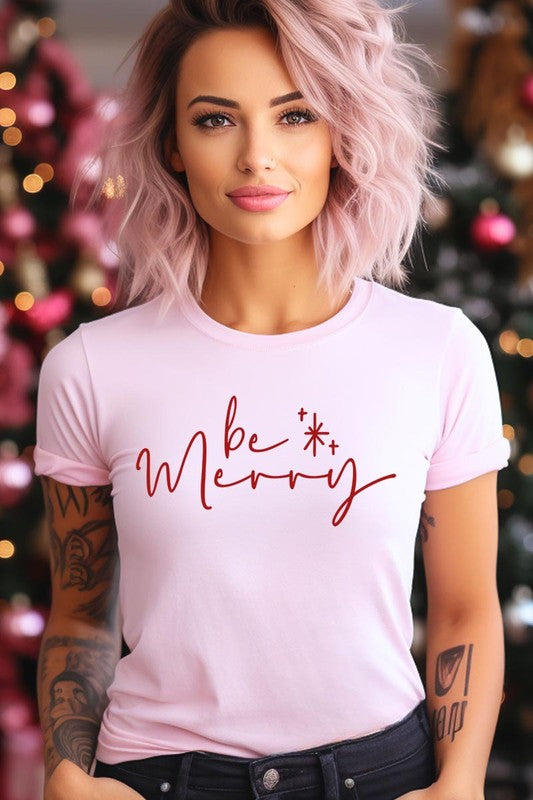 A person wearing the stylish and high-quality Be Merry Graphic Tee, featuring a decorative design with light-colored fabric and bold red text that reads "Be Merry." The unisex crew neck shirt is perfectly paired with them holding a red and black plaid garment.