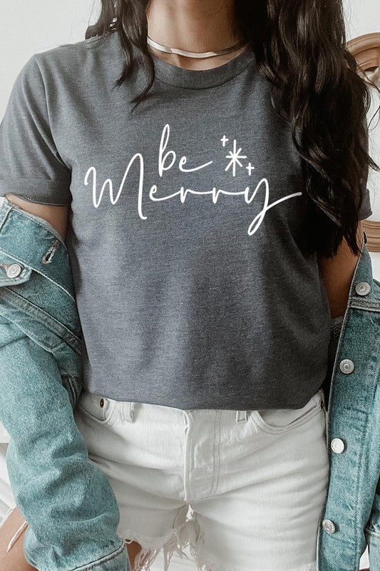 A person wearing the stylish and high-quality Be Merry Graphic Tee, featuring a decorative design with light-colored fabric and bold red text that reads "Be Merry." The unisex crew neck shirt is perfectly paired with them holding a red and black plaid garment.