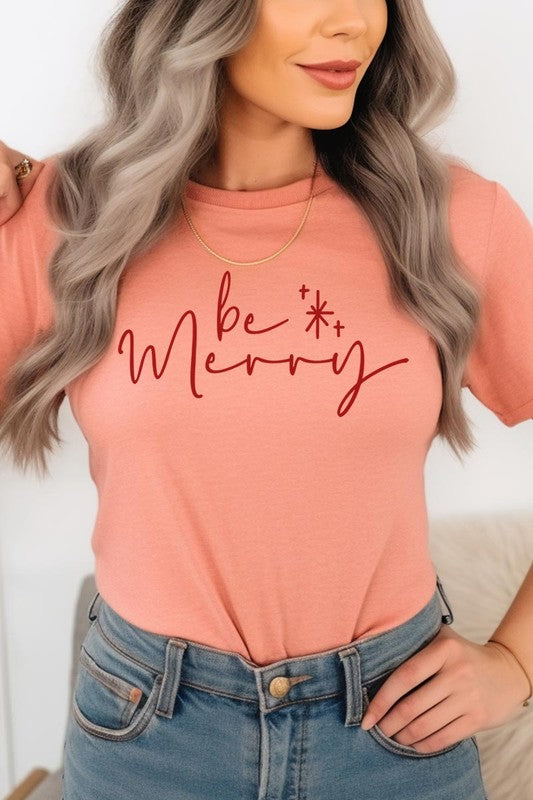 A person wearing the stylish and high-quality Be Merry Graphic Tee, featuring a decorative design with light-colored fabric and bold red text that reads "Be Merry." The unisex crew neck shirt is perfectly paired with them holding a red and black plaid garment.