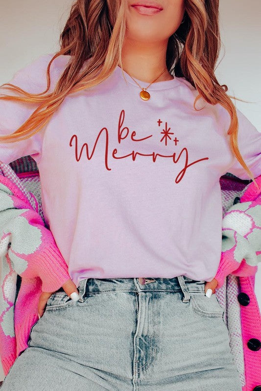 A person wearing the stylish and high-quality Be Merry Graphic Tee, featuring a decorative design with light-colored fabric and bold red text that reads "Be Merry." The unisex crew neck shirt is perfectly paired with them holding a red and black plaid garment.