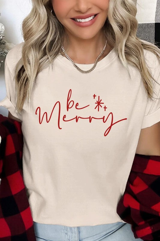 A person wearing the stylish and high-quality Be Merry Graphic Tee, featuring a decorative design with light-colored fabric and bold red text that reads "Be Merry." The unisex crew neck shirt is perfectly paired with them holding a red and black plaid garment.